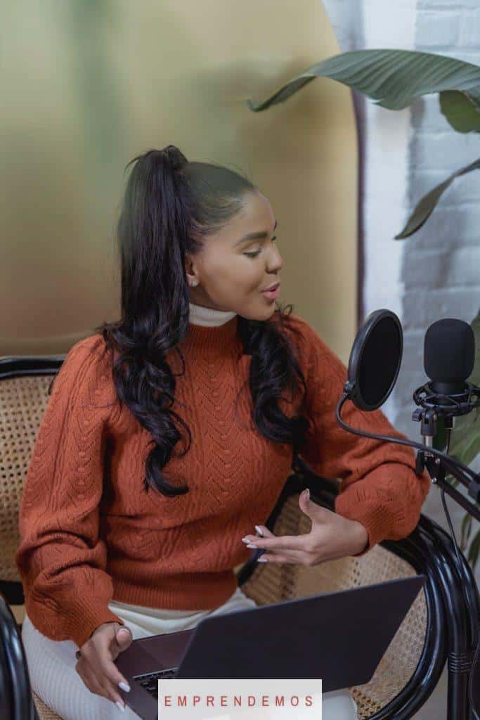 Confident young ethnic female blogger with long dark hair in casual clothes sitting on chair with laptop and speaking into microphone while recording podcast at home